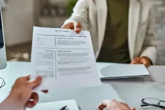 A person holds a resume in front of a computer, indicating a refresh of their CV for job applications.