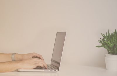 person writing on their laptop against white wall with green plant