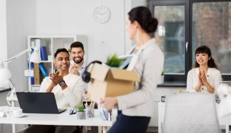 Woman carrying cardboard box through office while employees applaud 