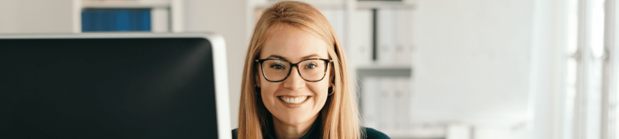 Blonde woman with glasses and long hair sat at a computer smiling to the front