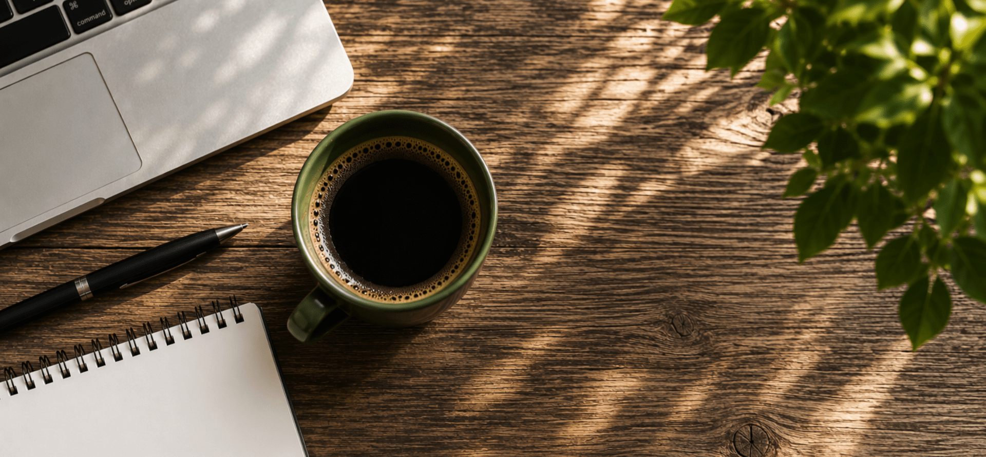 Sunlight coming through the window to a brown desk with a coffee, pad, pen and laptop.