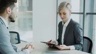 A man and woman sit at a table, focused on a tablet in front of them, engaged in discussion.