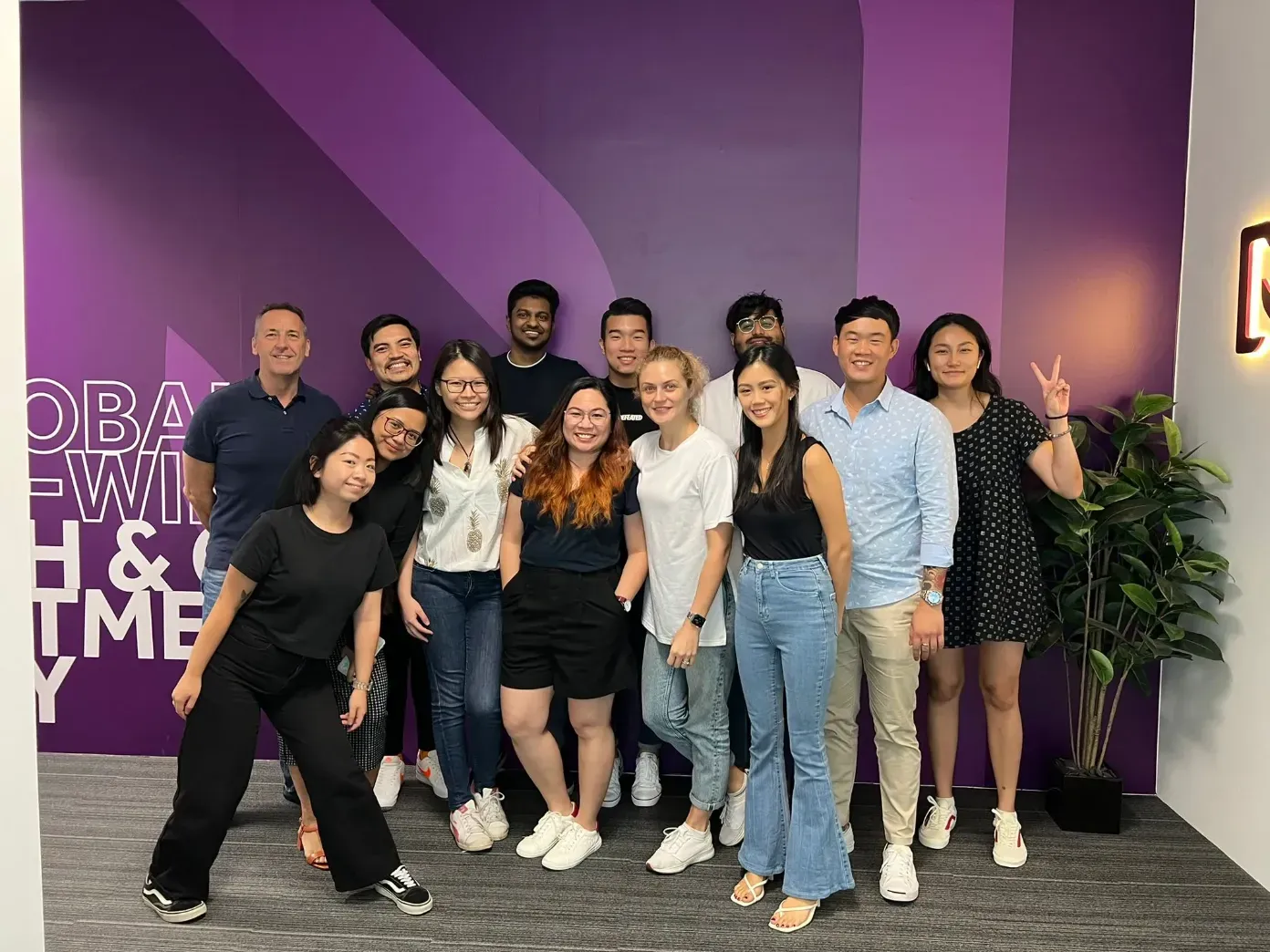 A group of smiling people standing in front of a purple wall dressed casually. 