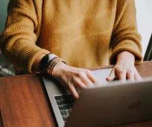 A woman focused on typing on her laptop, seated at a desk with a notepad beside her.