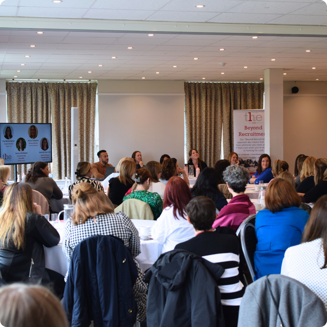 Group of people sat in chairs around circle tables listening to a panel of people speaking.