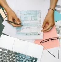 Hands holding a tax form over a cluttered desk with a laptop, eyeglasses, and euro bills, conveying a busy, focused atmosphere.
