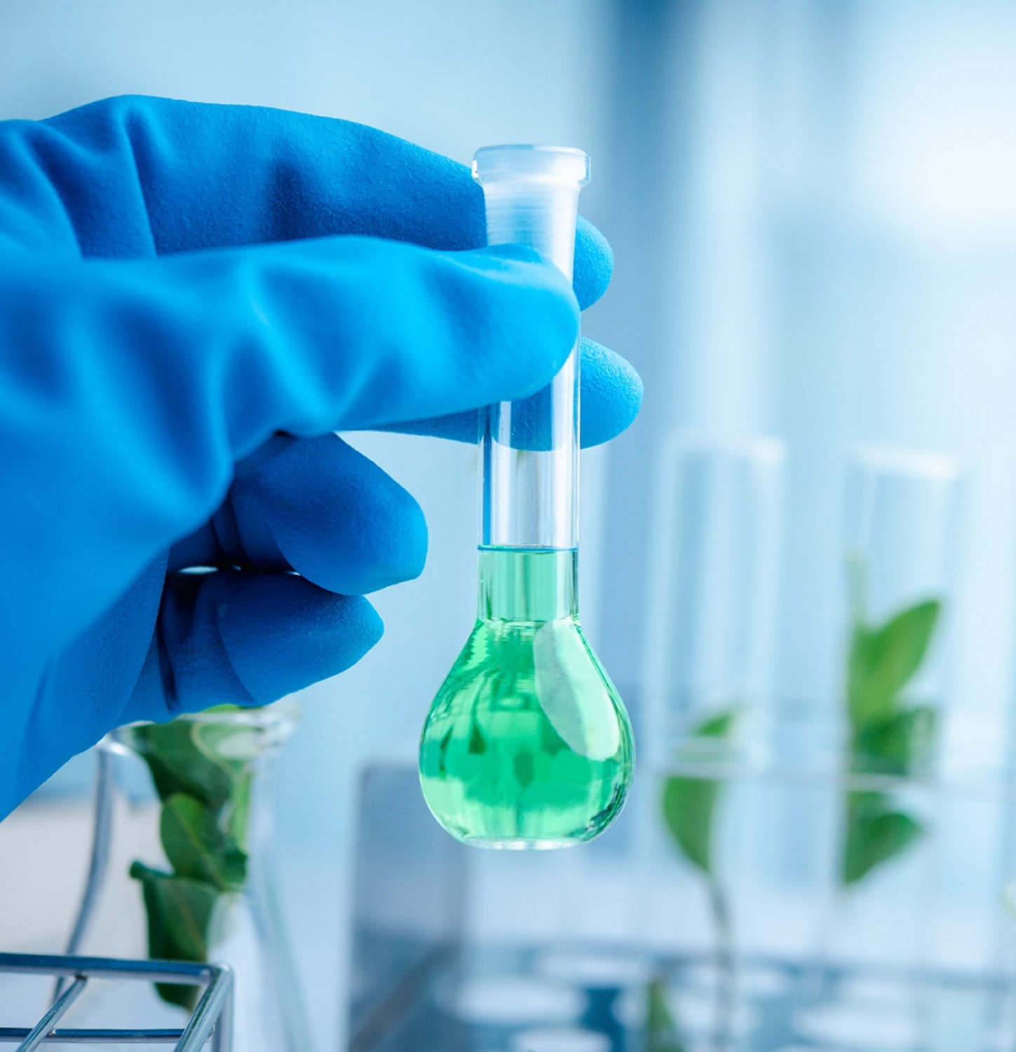 Zoom into a medical professional's hand. They are wearing a blue latex glove and holding a glass testing tube with a light green liquid inside.