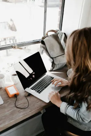A woman seated at a desk, focused on her laptop, with a notepad and pen beside her.
