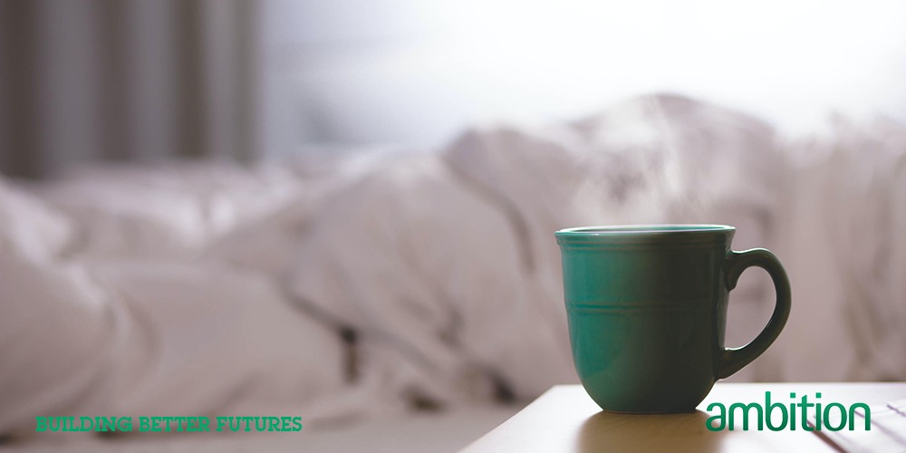 Green mug of tea on bedside table, person under duvet in the background; sick leave; day off sick