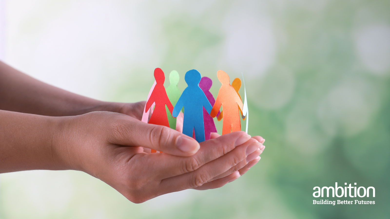 A pair of hands cupping and holding a group of different colour paper people, against a blurred green and white background 