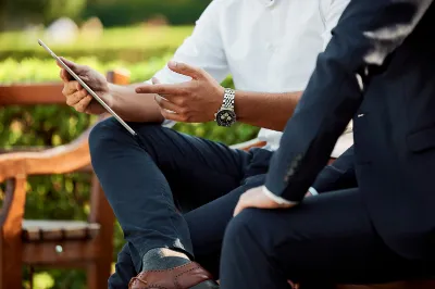 Two people in business attire sit outdoors on a bench, one holding a tablet and gesturing, suggesting a discussion or meeting.
