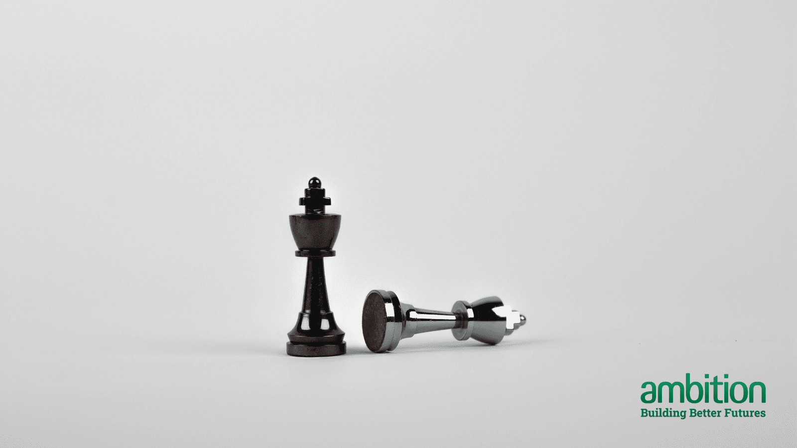 Light grey background with a black king chess piece in the middle and a silver king chess piece laying on it's side next to it