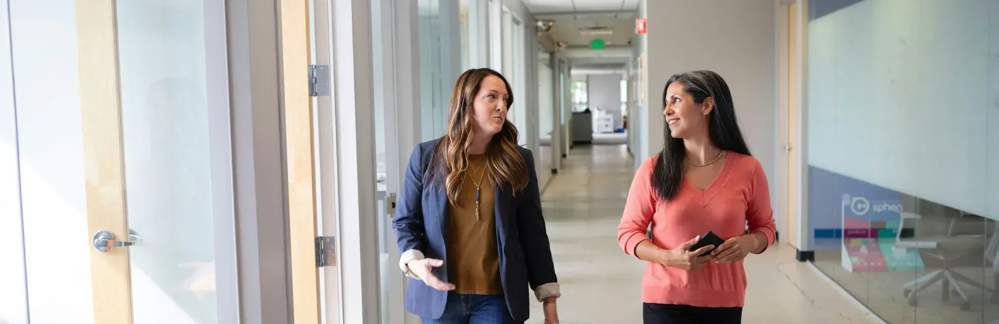 Two women walking and talking in a modern office hallway with glass doors and bright lighting.