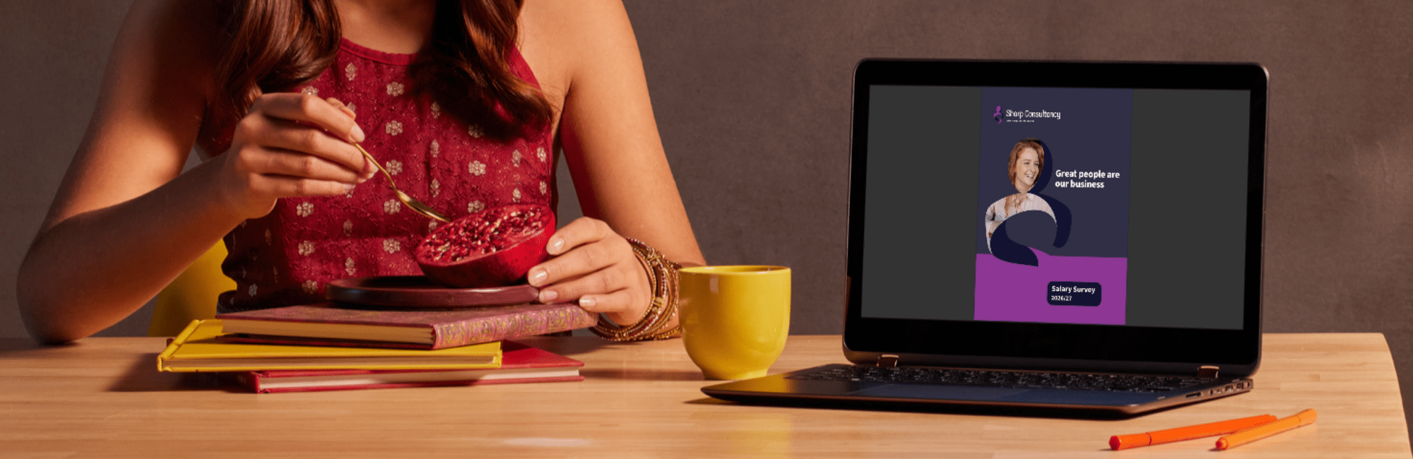 A woman sits at a table with a laptop and a cup of coffee, preparing to download the Sharp Consultancy 2026/27 salary survey.