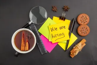 Sticky notes, a coffee cup, cinnamon, and office supplies arranged on a black background with a "We Are Hiring" message.