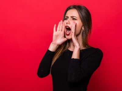 Lady against red wall holding hand up to mouth and shouting 