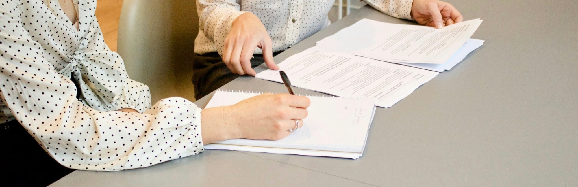 Two people in polka dot shirts sit at a table, reviewing documents with focus and collaboration. One writes in a notebook while the other gestures.