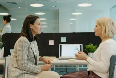  Two women engaged in conversation at a desk in a professional office environment.
