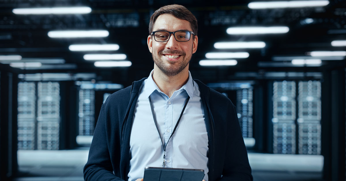 White Male It Specialist Standing With A Tablet In A Dark Data Center With Many Servers