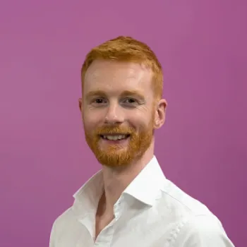 A man with red hair and a beard, wearing a white shirt, smiles in front of a plain purple background