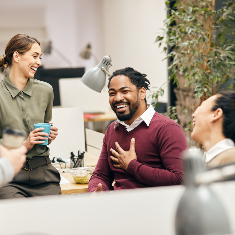 A diverse group of colleagues in an office setting, gathered around a workspace and laughing together. One man with a beard and wearing a maroon sweater is holding his chest while smiling, while the others are holding coffee cups and engaged in a lively conversation. The office environment is bright and modern, with plants and desks in the background.