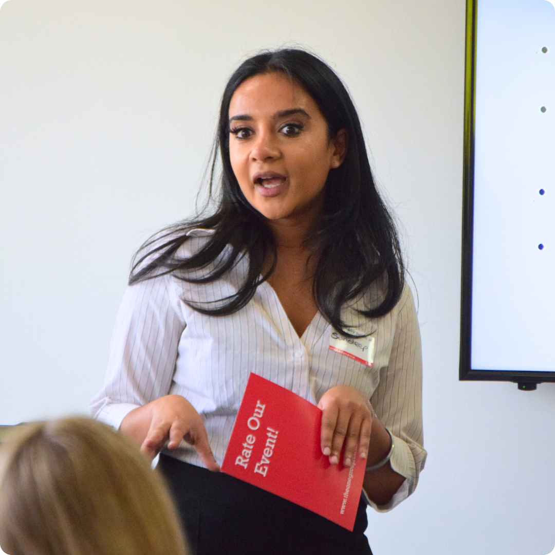 Woman stood talking to an audience at an event, holding a red card saying 'rate our event'.