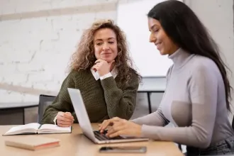 Two women engaged in a mentor-mentee discussion at a table with a laptop between them.