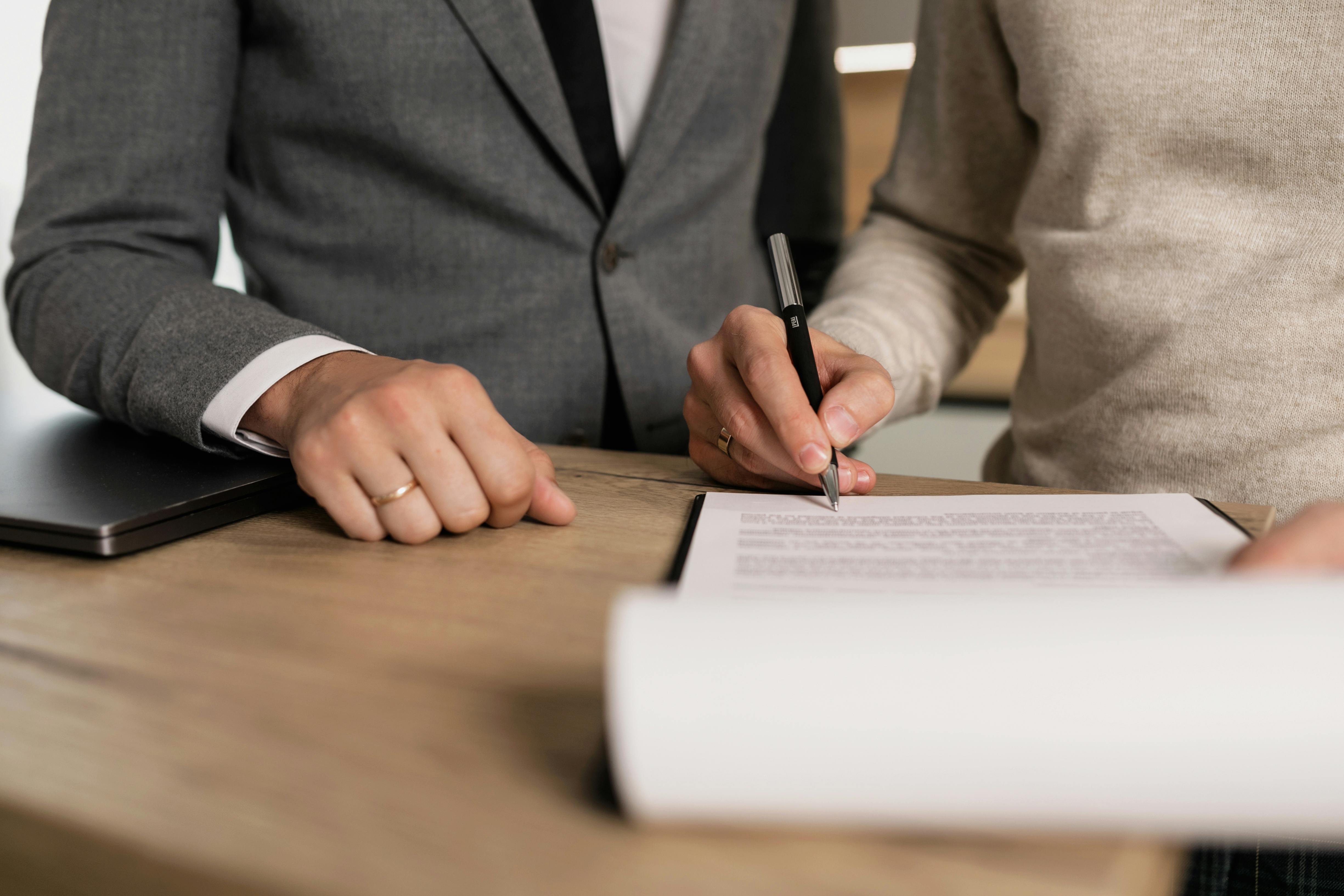 Two individuals in business attire focus on signing a document at a wooden desk. One holds a pen, the other has hands resting on a closed laptop.