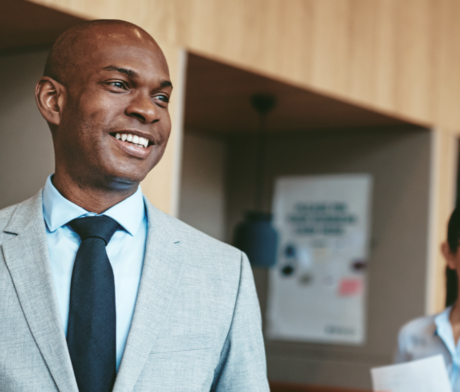 A smiling man in a light grey suit, light blue shirt, and dark tie standing in a professional office environment. The background is slightly blurred, showing a modern office setting with a wall poster and another person partially visible.