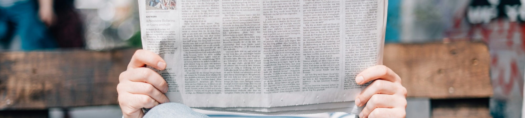 A person sits on a bench, engrossed in reading a newspaper on a sunny day in a park.