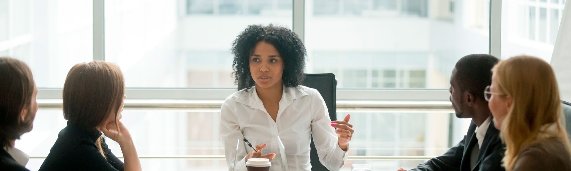 A young professional woman in a white shirt addresses four smartly dressed colleagues during a business meeting