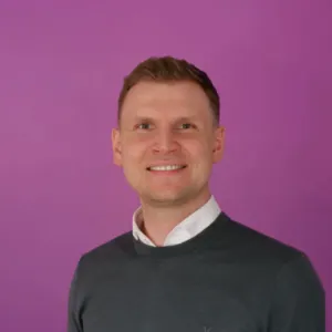 A man wearing a green sweater stands against a vibrant purple wall, creating a striking color contrast.