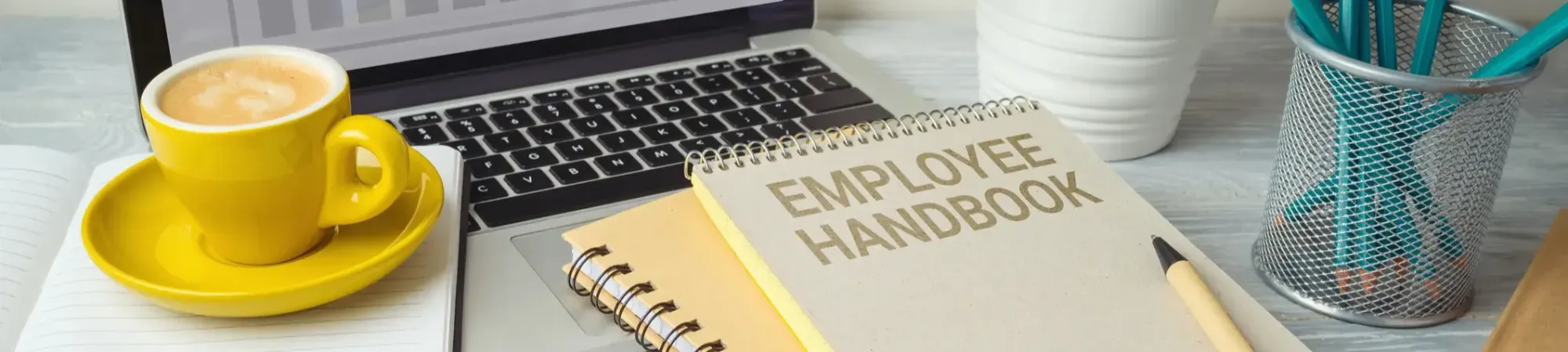 Een desktop met een laptop, een boekje waar op staat "employee Handbook" en een kopje koffie