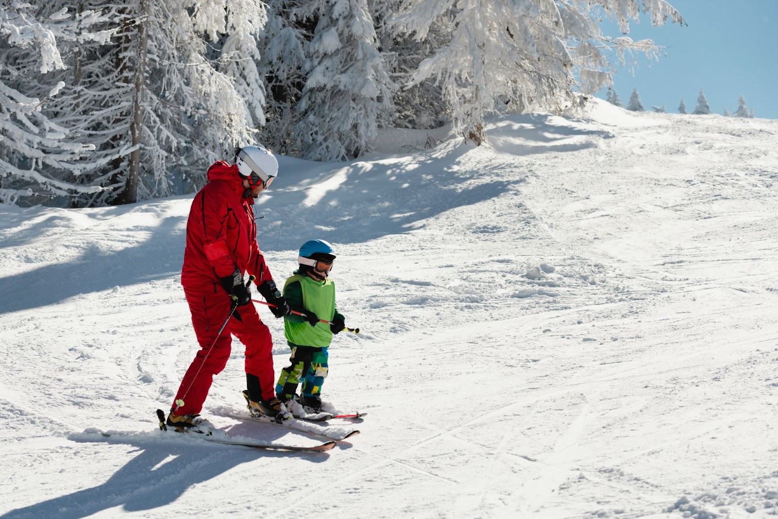 Nanny and child skiing down slope under blue sky.