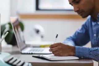 A man sits at a desk, writing in a notebook with a focused expression.