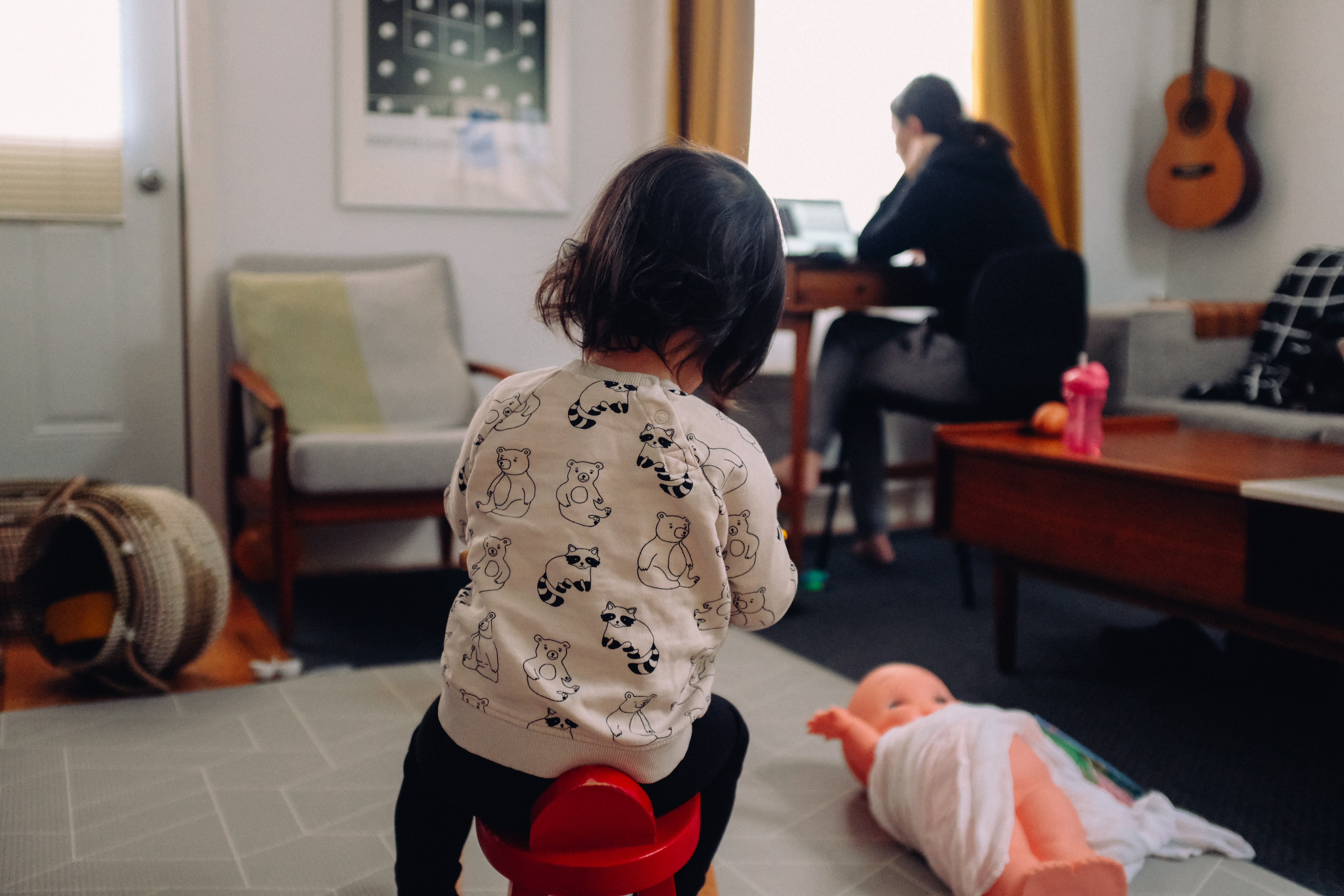 In a living room, a young girl uses a potty as her parent, an accountant, works from home, balancing care and work.