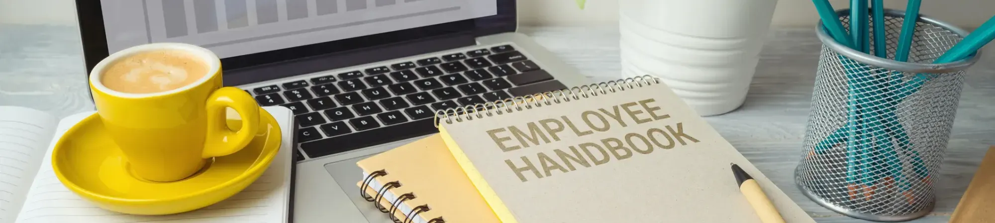 a Desktop with a laptop, and a little book which says "employee handbook" and a cup of coffee