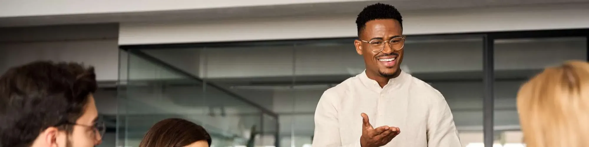 A professional setting where a man with glasses is standing and smiling while speaking to a group. He is casually dressed in a light-coloured shirt, engaging confidently with the other seated individuals, who are visible only from the back and side. The environment appears to be a modern office or meeting room with glass walls.