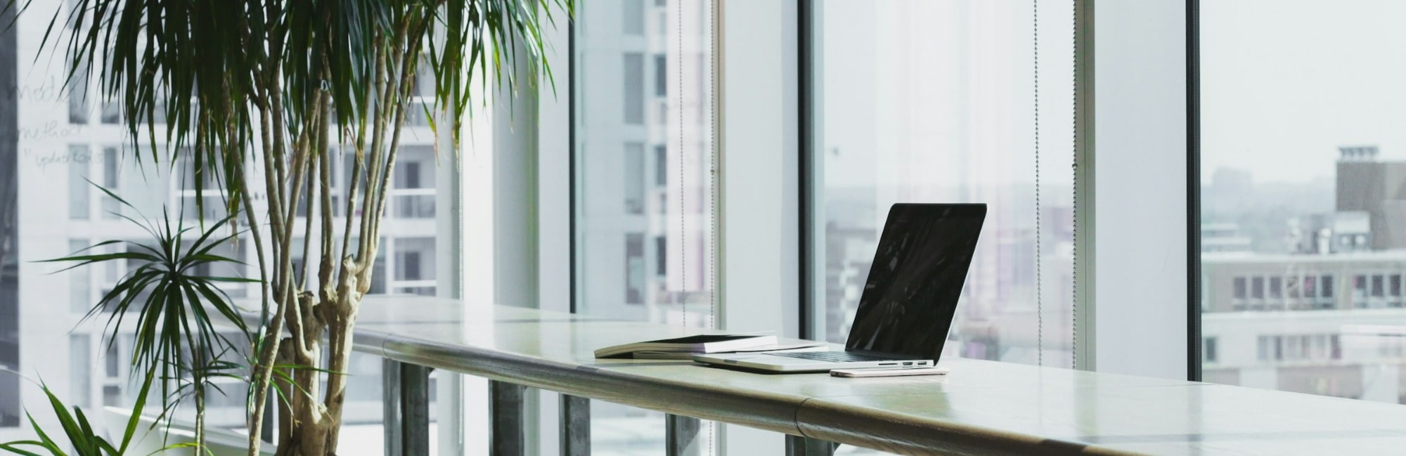 A laptop sits on a table in front of a large window, allowing natural light to illuminate the workspace.