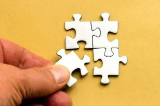 A hand grips a white puzzle piece on a vibrant yellow background, representing the concept of the right fit.