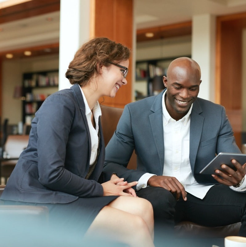 Two professionals sitting together in a modern office setting. A man in a dark suit jacket and white shirt is showing a tablet to a woman in glasses and a navy suit. Both are smiling and engaged in a conversation.