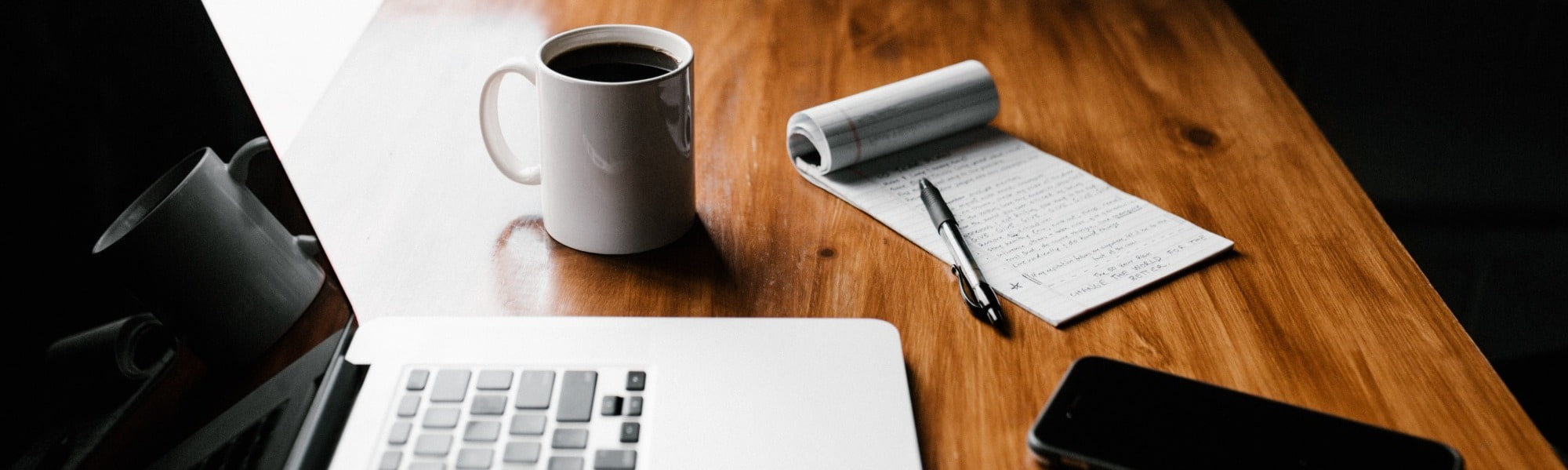 A lap top sits open on a desk alongside a mug and notebook and pen