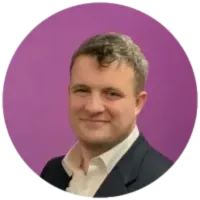A man in a suit and tie stands confidently in front of a vibrant purple wall.