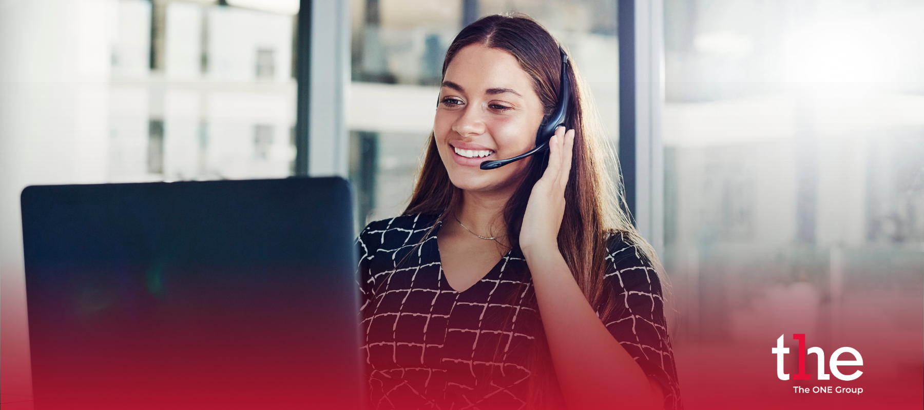 Woman on phone with a headset looking at her computer in an office meeting room environment. Image has a red gradient at the bottom with The ONE Group's logo in the right bottom corner.