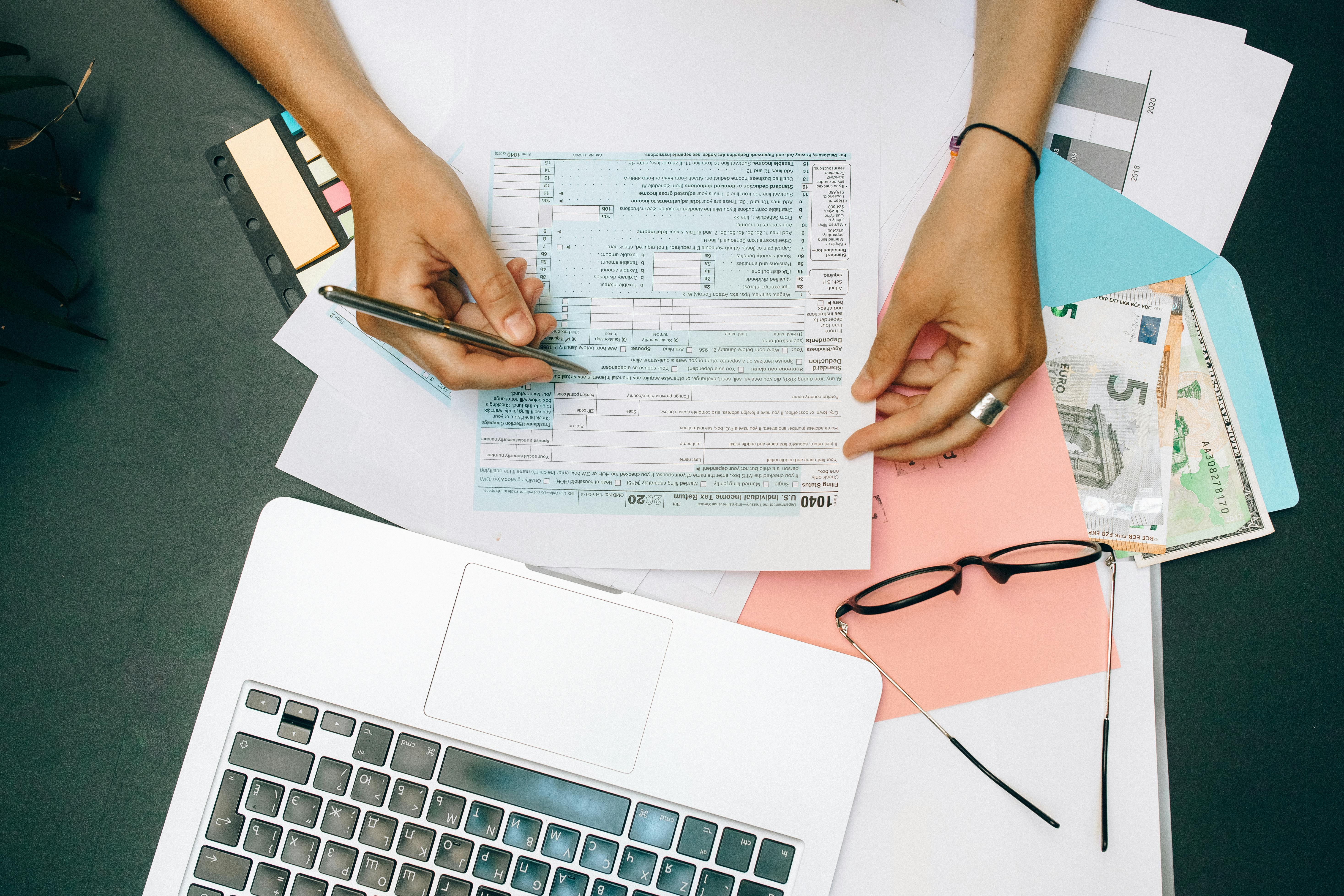 Hands holding a tax form over a cluttered desk with a laptop, eyeglasses, and euro bills, conveying a busy, focused atmosphere.