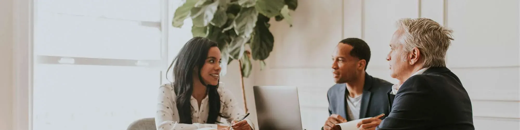 Three professionals engaged in a discussion around a table in a bright office setting. A woman is smiling and holding a pen, while two men listen attentively. A laptop is placed on the table, and a large plant decorates the background.