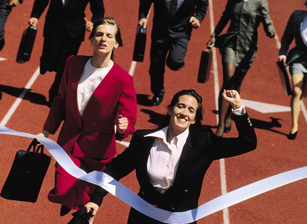 Women in business attire running a race on a race track and crossing the finish line